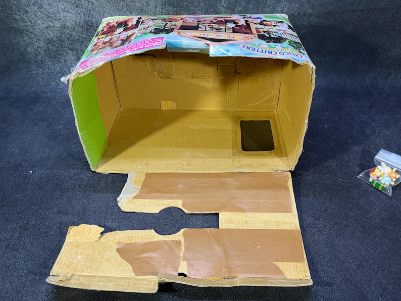 May include: An open, partially assembled cardboard box with a colorful printed top. The box is tan with green and brown accents. A small plastic bag of colorful beads sits to the right of the box.
