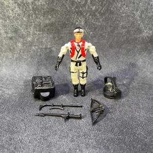 Puede incluir: Figura de acción vintage de un guerrero ninja con uniforme blanco y rojo, guantes y botas negros. Accesorios: mochila negra, carcaj, arco y dos armas. La figura tiene una cinta blanca con un símbolo rojo.