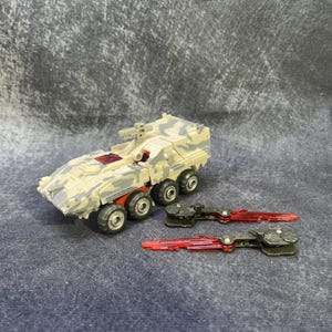 Puede incluir: Un vehículo militar de juguete con pintura de camuflaje y cuchillas de plástico rojo. El vehículo tiene cuatro ruedas y una torreta de cañón en la parte superior.