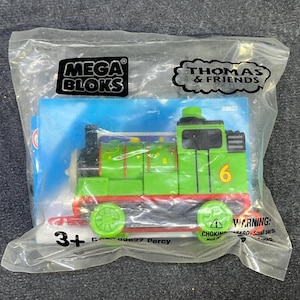 May include: A green and red plastic toy train engine with the number 6 on the side. The engine is labelled "Percy" and is part of the Thomas & Friends Mega Bloks range. The packaging is for ages 3 and up.