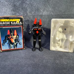 May include: A vintage Baron Karza action figure with its original box and packaging. The black and red figure has a detailed design, and the box features the figure's image and text. The packaging includes a white molded insert.