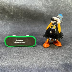 May include: A small figurine of a character with a black cape, orange feet, and yellow arms, holding a blue and white object. Beside it is a miniature skateboard with green edges and the text "World Industries".