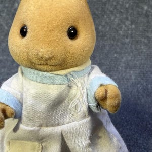 May include: A tan-colored rabbit figurine wearing a white coat with light blue trim. The figurine has black eyes and a small, stitched mouth. The coat has a small tear on the front. The toy is a collectible item.