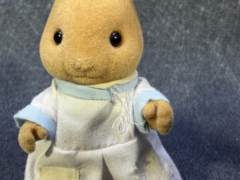 May include: A tan-colored rabbit figurine wearing a white coat with light blue trim. The figurine has black eyes and a small, stitched mouth. The coat has a small tear on the front. The toy is a collectible item.