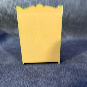 Op de afbeelding: Een kleine, gele houten kast met een geschulpte bovenkant en groene rand. De kast is rechthoekig en lijkt een miniatuur decoratief stuk te zijn. De achtergrond is donkerblauw.