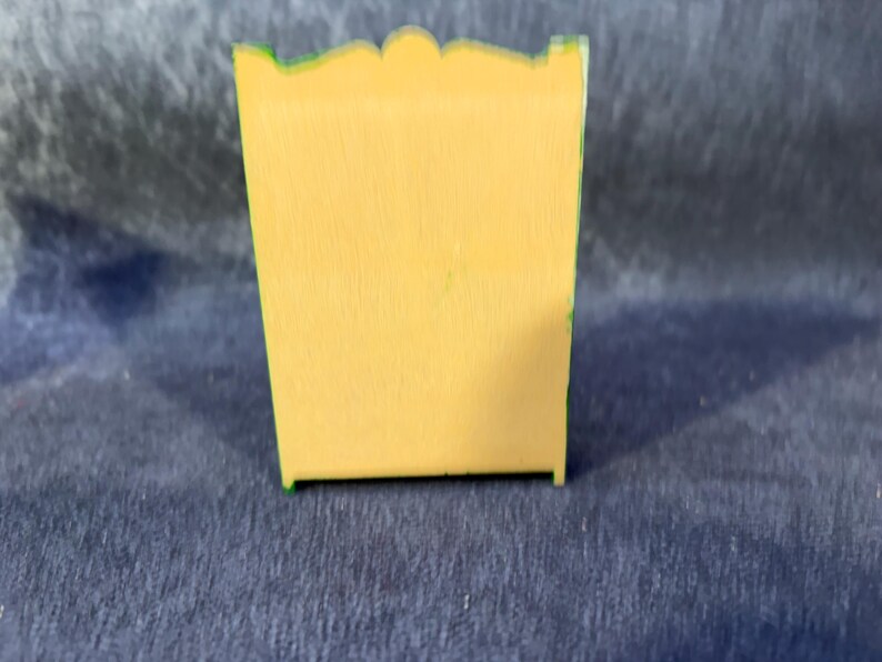 Op de afbeelding: Een kleine, gele houten kast met een geschulpte bovenkant en groene rand. De kast is rechthoekig en lijkt een miniatuur decoratief stuk te zijn. De achtergrond is donkerblauw.