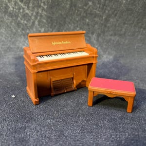 May include: A miniature brown piano with white keys and a matching red-topped bench. The piano has the text "Sylvanian families" on the top. The miniature piano is a toy.