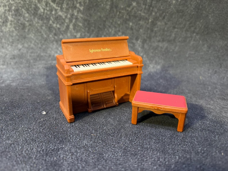 May include: A miniature brown piano with white keys and a matching red-topped bench. The piano has the text "Sylvanian families" on the top. The miniature piano is a toy.