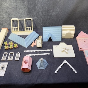 Könnte beinhalten: Eine Sammlung von Puppenhaus-Komponenten in verschiedenen Farben, darunter eine hellrosa Hauswand, blaue Dachabschnitte und cremefarbene architektonische Elemente. Das Set enthält Fenster, Türen und dekorative Zierleisten, ideal für ein Miniatur-Bauprojekt.