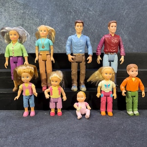 Può includere: Una collezione di nove bambole in plastica di varie età, tra cui due figure adulte con camicie e pantaloni, due figure adulte con capelli grigi o biondi e cinque figure più piccole di bambini e neonati che indossano abiti colorati.