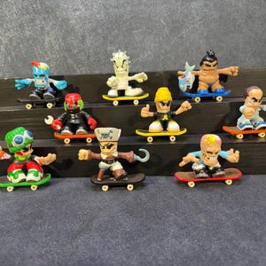 Pode incluir: Uma coleção de dez pequenas figuras de personagens estilizados, cada uma montada num skate de plástico em miniatura. As figuras apresentam designs diversos, incluindo um pirata com um gancho, uma múmia, um lutador de sumo e várias criaturas coloridas.