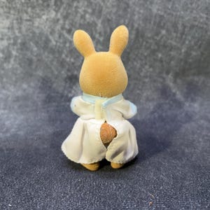 May include: A small, tan rabbit figurine wearing a white and light blue outfit. The rabbit has long ears and is facing away from the camera. The outfit includes a collar and a skirt-like bottom.