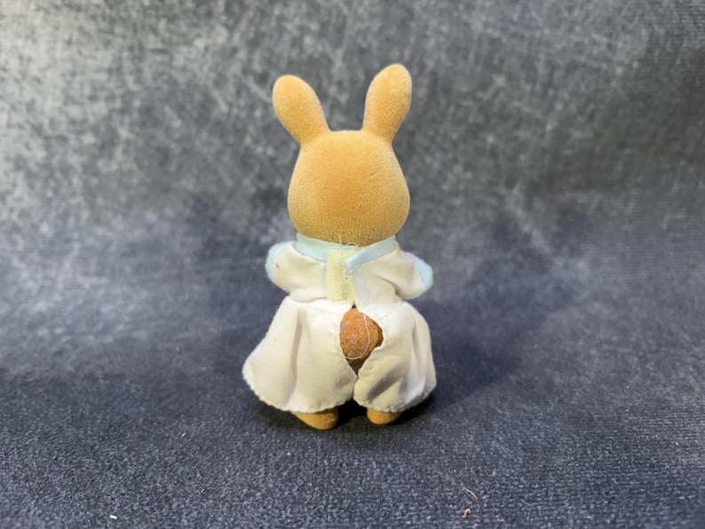 May include: A small, tan rabbit figurine wearing a white and light blue outfit. The rabbit has long ears and is facing away from the camera. The outfit includes a collar and a skirt-like bottom.