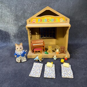 May include: A miniature preschool playset with a building, piano, and several animal figurines. The building has a sign that reads "Preschool". There are also three miniature beds with floral patterned sheets.