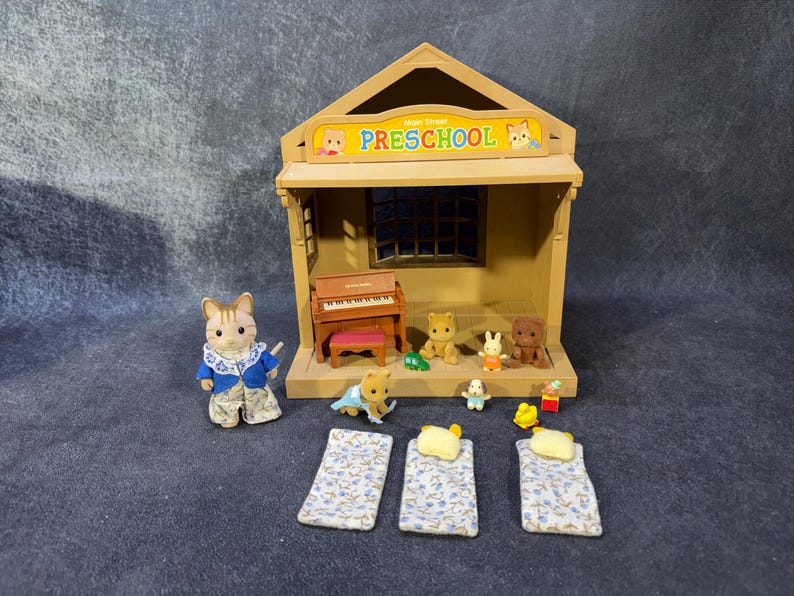 May include: A miniature preschool playset with a building, piano, and several animal figurines. The building has a sign that reads "Preschool". There are also three miniature beds with floral patterned sheets.