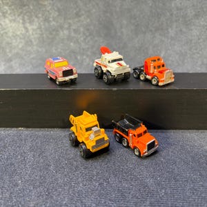 以下が含まれることがあります： ピンクのSUV、赤いクレーン付きの白いトラック、オレンジ色のセミトラック、黄色のダンプカー、黒い荷台を持つオレンジ色のダンプカーの計5台のミニチュアダイキャスト製おもちゃの乗り物。すべて黒いプラスチック製の車輪が付いています。