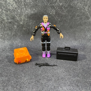 May include: A vintage action figure of a man in a black and orange jumpsuit with a pink vest and boots. Accessories include an orange box, a black briefcase, and a black rifle. The figure has gray hair.