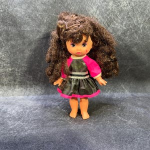 Può includere: Una piccola bambola con capelli castani ricci, che indossa un vestito bianco e nero con una giacca rosa. La bambola ha un viso dipinto con occhi marroni e un blush rosa.