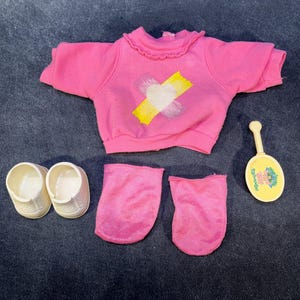 May include: A pink doll outfit set, including a short-sleeved top with a white heart and yellow cross design, matching socks, white shoes, and a yellow rattle. The items are arranged on a dark blue surface.