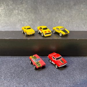 以下が含まれることがあります： 5台のミニチュア玩具車のコレクション。3台の黄色い車が並んでおり、黒い炎やストライプの模様が施されています。手前には2台の赤い車があり、1台は黒と黄色の動物柄、もう1台は黒いレーシングストライプが特徴です。