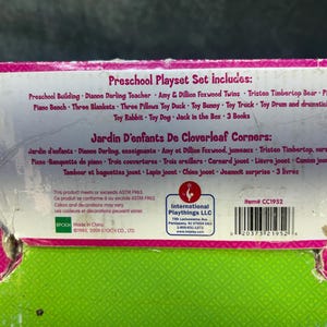 May include: A close-up of a preschool playset box with text in English and French. The box lists the included items: a preschool building, teacher, twins, bear, piano, blankets, pillows, and toys. The box is pink, green, and white.