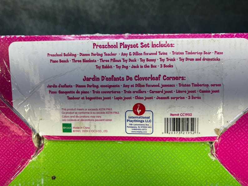 May include: A close-up of a preschool playset box with text in English and French. The box lists the included items: a preschool building, teacher, twins, bear, piano, blankets, pillows, and toys. The box is pink, green, and white.