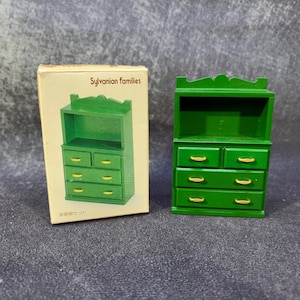 Op de afbeelding: Een groene miniatuur houten kast met lades en een bovenste plank, naast de originele doos. De doos heeft de tekst "Sylvanian Families" en een illustratie van de kast. De kast heeft goudkleurige ladeknopen.