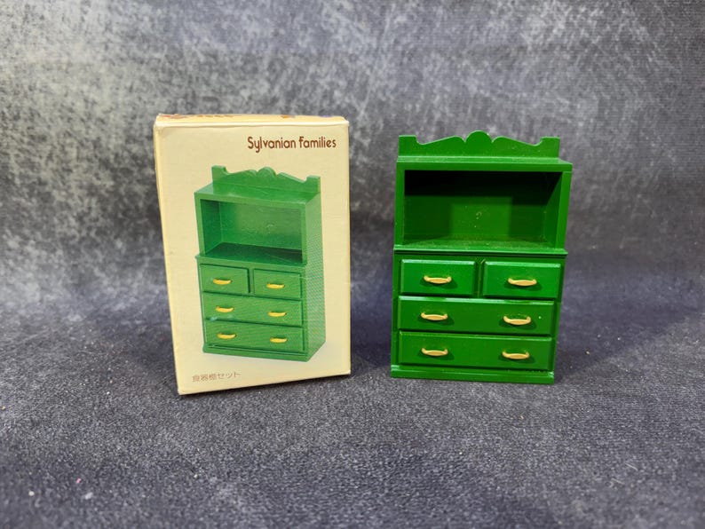 Op de afbeelding: Een groene miniatuur houten kast met lades en een bovenste plank, naast de originele doos. De doos heeft de tekst "Sylvanian Families" en een illustratie van de kast. De kast heeft goudkleurige ladeknopen.