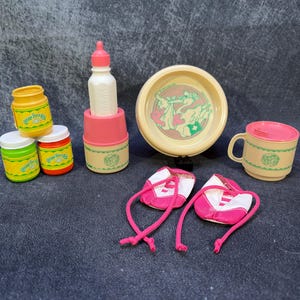 May include: A selection of vintage baby doll accessories in pastel shades. Includes a baby bottle, a bowl, a cup, small jars, and a pair of pink and white shoes. The jars have the text "Baby Twinkle".