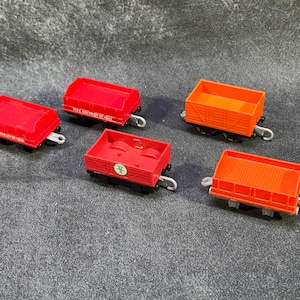 Op de afbeelding: Vijf speelgoedtreinwagons in rood en oranje. De wagons hebben zwarte wielen en zilveren haken. Sommige wagons hebben open bovenkanten, andere dichte. Eén rode wagon heeft witte tekst.