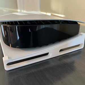 May include: A white stand with a black PlayStation 5 console on top. The stand has a curved design and is made of plastic. The console is sitting on the stand with the disc drive facing up.