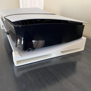 Puede incluir: Una consola de videojuegos blanca y negra sobre un soporte blanco. La consola tiene una parte superior negra y una inferior blanca. El soporte está hecho de plástico y tiene un diseño curvo.