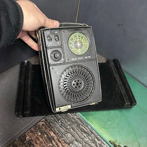 Könnte beinhalten: Ein schwarzes tragbares AM/FM-Radio mit einer runden grünen Senderskala, einem Lautstärkeregler und einem großen Lautsprechergitter. Auf der Vorderseite befindet sich der Schriftzug SOLID STATE AM/FM sowie ein kleines rechteckiges Etikett.