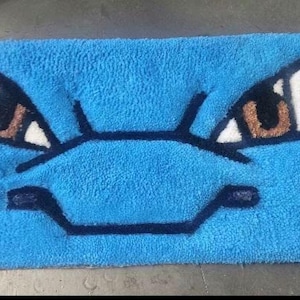 May include: A rectangular blue rug featuring a cartoon face with brown eyes, white accents, and black outlines. The rug has a plush texture and is likely made of a soft material. The design is reminiscent of a popular character.