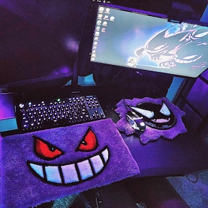 Könnte beinhalten: Ein Gaming-Setup mit einer schwarzen Tastatur, einer Maus und zwei lila Teppichen mit einem Cartoon-Gesicht-Design. Ein Computermonitor zeigt ein Cartoon-Bild an. Das Setup ist mit lila Licht beleuchtet.