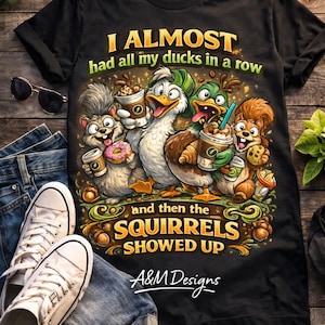 May include: Black t-shirt with a humorous graphic featuring cartoon ducks and squirrels holding coffee cups and donuts. The text reads "I ALMOST had all my ducks in a row...and then the SQUIRRELS SHOWED UP." The shirt is displayed with jeans and sneakers.