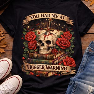 Puede incluir: Camiseta negra con una calavera atravesada por una espada, rodeada de rosas rojas y libros. La camiseta tiene el texto "YOU HAD ME AT" encima de la calavera y "TRIGGER WARNING" debajo. Se ven zapatillas blancas y vaqueros azules.