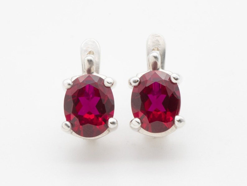 May include: A pair of silver earrings with red oval-shaped gemstones. The earrings have a clip-on closure.
