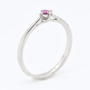 Puede incluir: Un anillo de banda de plata simple con una sola piedra preciosa de rub&iacute; rosa engastada en el centro.