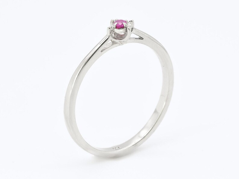 Puede incluir: Un anillo de banda de plata simple con una sola piedra preciosa de rub&iacute; rosa engastada en el centro.