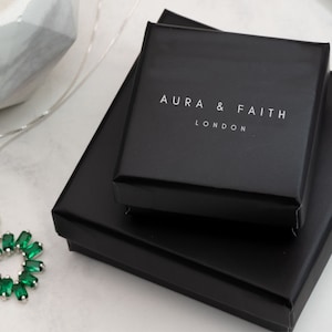 Puede incluir: Un collar de plata con un colgante en forma de coraz&oacute;n verde. El collar est&aacute; empaquetado en una caja negra con las palabras "Aura & Faith London" impresas en la parte superior.