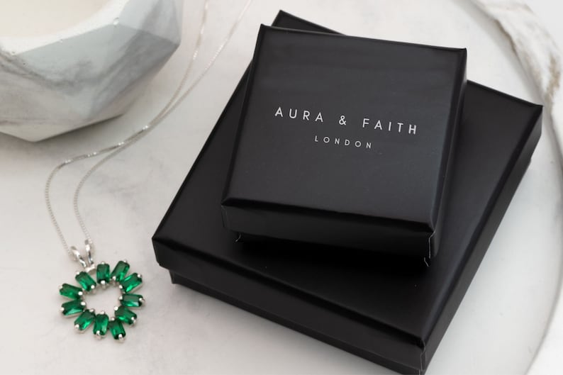 Puede incluir: Un collar de plata con un colgante en forma de coraz&oacute;n verde. El collar est&aacute; empaquetado en una caja negra con las palabras "Aura & Faith London" impresas en la parte superior.