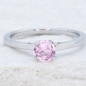 May include: A silver ring featuring a round, pale pink gemstone. The ring has a minimalist design with a slender band and a four-prong setting. The ring is displayed against a white backdrop.