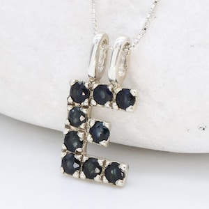 Op de afbeelding: Zilveren ketting met hanger in de vorm van de letter E, bezet met kleine blauwe edelstenen.