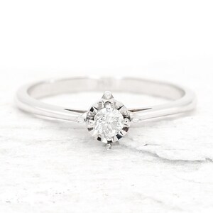 May include: A simple solitaire engagement ring with a single round diamond set in a white gold band.