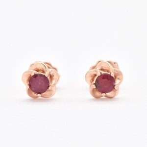 May include: A pair of rose gold stud earrings with a red ruby gemstone in the centre of each flower-shaped setting.
