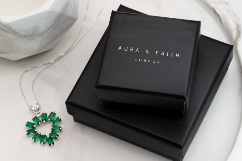 Puede incluir: Un collar de plata con un colgante en forma de coraz&oacute;n verde. El collar est&aacute; empaquetado en una caja negra con las palabras "Aura & Faith London" impresas en la parte superior.
