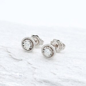 May include: A pair of stud earrings featuring a central, diamond-like stone. The earrings are set in a silver-coloured metal with a textured, rope-like border. The earrings are presented on a white, textured surface.