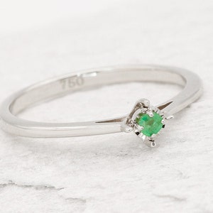 May include: A delicate silver ring with a small, green emerald gemstone set in a prong setting. The ring is marked "750" on the inside band.