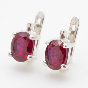May include: A pair of silver earrings with red oval-shaped gemstones. The earrings have a simple design with a small loop for attaching to the ear.